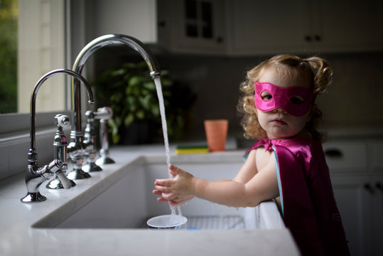 Portrait of girl in superhero costume washing hands in kitchen sink