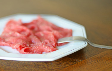 Beef carpaccio with olive oil and fresh ground white pepper on white porcelain plate with silver lining and with stainless steel fork, brown oak wooden background. Very shallow depth of field