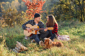 Happy caucasian couple with guitar and their dog having rest on picnic in the park on the lawn.