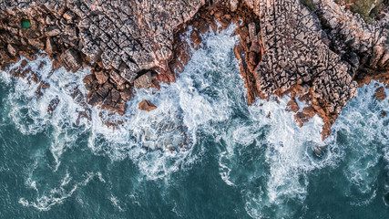 Drone aerial top view of sea waves hitting rocks on the beach with turquoise sea water. Amazing rock cliff seascape in the Portuguese coastline, Cascais.