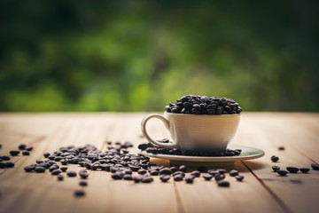 White cup full of coffee beans on Roasted Coffee Beans and wooden table in nature background