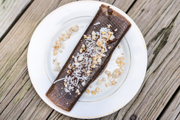 Flat top closeup of chocolate crepe dessert wrap with rice crisps and coconut flakes made in dehydrator with bananas on plate wooden table