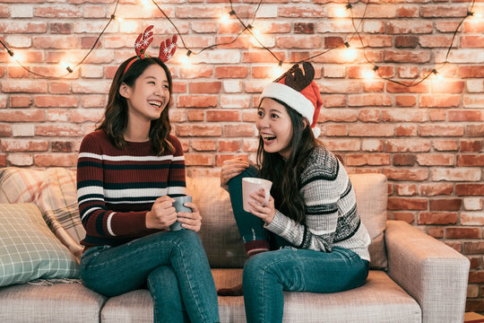 Cheerful Asian Female Friends Having Fun Laughing While Chatting Sit In Sofa. Young Girls Chill Out On Christmas Eve At Home Celebrated Together. Two Elegant Women In Sweater Holding Hot Chocolate