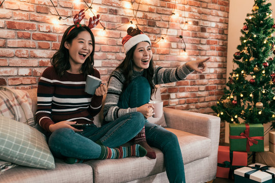 asian female friends watching movie and having fun at Christmas eve sitting on couch with gift boxes under xmas tree. young girls cheerfully point to television screen comedy showing sharing laughing
