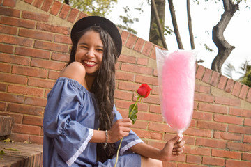 A young, beautiful and in love young girl with a dress of bare shoulders and a fedora hat just received a red rose and a cotton candy. She is very happy and radiant with joy.