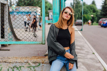 Fashion portrait of trendy young woman wearing sunglasses, jeans with halls and bomber jacket in...