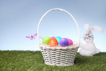 Little white rabbit close to a wicker basket full of colorful Easter eggs