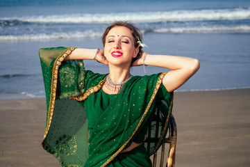 happy beautiful indian bindi woman in green traditional sari sitting on a chair on beach.female sunbathing enjoying summer vacation on the ocean spf sunscreen