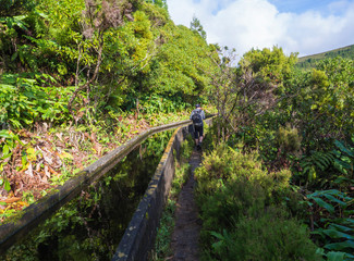 Fototapeta premium Men hiker walking on footpath along Lavada - water irrigation canals covered by moss and lush vegetation on hiking trail Praia Lagoa de Fogo, Sao Miguel island, Azores, Portugal