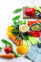 Plate of fresh organic vegetables salad with humus and green pesto dip on white marble background, healthy flat layout food concept with fresh spring or summer vegetables, light dinner