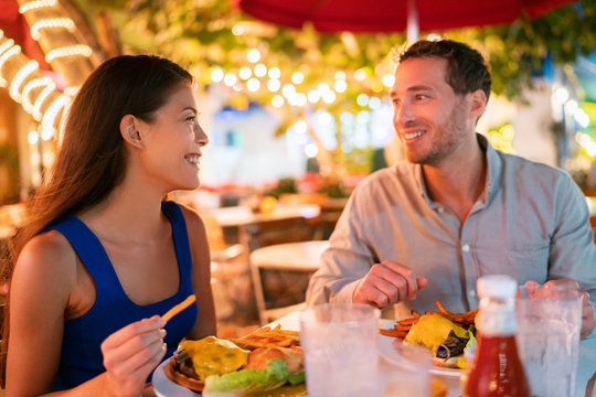 Couple Eating Hamburgers At Outdoor Restaurant Terrace Happy Tourists On Summer Vacation. Florida Travel People Eating Food At Night During Holidays In Miami. Asian Caucasian Interracial Young Adults.