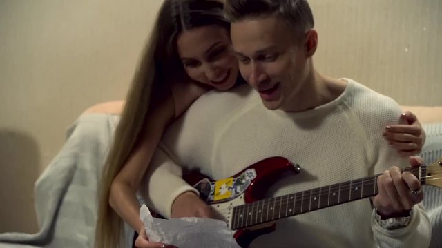 Man Sitting On A Couch, Playing Guitar With A Hug From His Girlfriend. Young Man Is Playing The Electric Guitar, Young Pretty Woman Is Sitting Behind Him, Hugging Him, Singing.