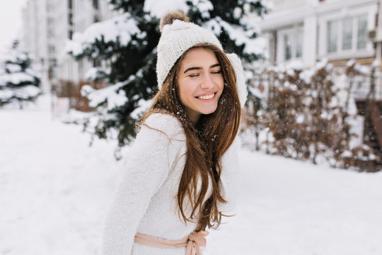 Outdoor portrait of fascinating brunette lady posing with romantic smile on snowy background. Photo of pretty laughing girl in knitted hat and soft sweater on the winter street..