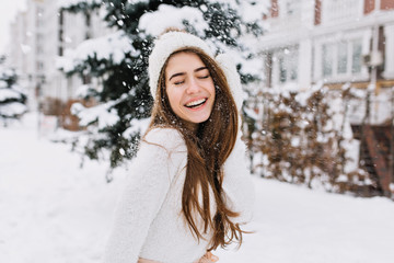 Happy winter moments of joyful young woman with long brunette hair, white winter clothes having fun on street in snowing time. Expressing positivity, true brightful emotions, smiling with closed eyes