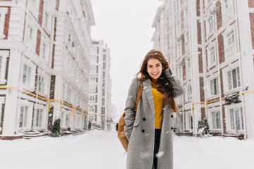 Charming young woman in coat with long brunette hair enjoying snowfall in big city. Cheerful...