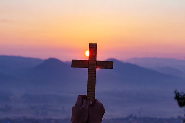 human hands praying to the GOD while holding a crucifix symbol with bright sunbeam on mountain at sunset