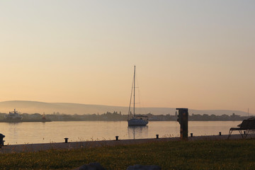 The sailing yacht enters under the motor in the marina in the rays of the morning sun. Dawn in the port. Empty bench on the pier against the lighthouse. Symbols of return and waiting. A calm romantic 