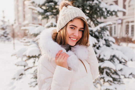 Romantic young woman in trendy knitted hat posing with smile standing near christmas snowy tree. Outdoor photo of laughing european woman wears ring walking down the street in winter day.