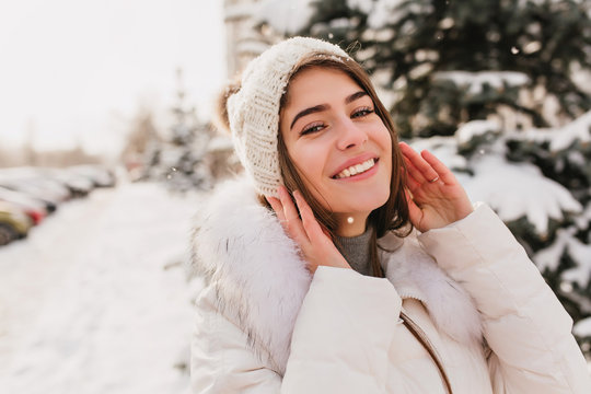 Close-up portrait of gorgeous girl with blue eyes posing on the street in snowy winter day. Outdoor photo of charming female model in knitted hat laughing on blur city background in cold morning..