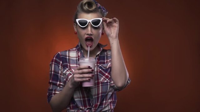 Funny blonde girl with retro hairstyle is drinking a milkshake and crossing her eyes, slow motion
