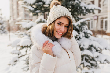 Fashionable young woman in white knitted hat smiling friendly to camera on street full with snow. Amazing european girl enjoying winter time on snowy tree background. .