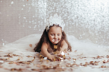 little girl in white dress lies on the floor and looks away on background with silver bokeh.