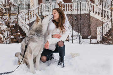 Pretty caucasian lady in white jacket kissing her husky dog during walk in winter day. Attractive young woman wears ripped jeans playing with pet in snowy morning..
