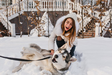Chilling girl with long shiny hair having fun with husky in frosty december day. Outdoor photo of smiling young lady relaxing with her dog during winter holidays.