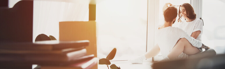 selective focus of beautiful young couple hugging and sitting on window sill at home with copy space and sunlight