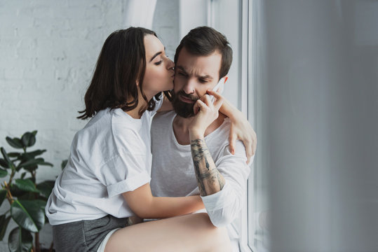 beautiful woman kissing man talking on smartphone at home