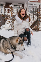 Husky dog lying on snow while long-haired girl laughing on background. Inspired european young lady in ripped pants spending time outdoor with her pet in cold day.