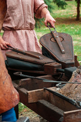 Blacksmith in ancient clothes using pair of bellows to light fire in furnace in medieval smithy....