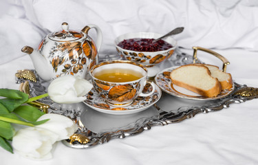 mug of tea, 3 white tulips, kettle, slices of bread, jam, tea set on a metal tray on a white sheet