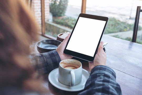 Mockup image of woman's hands holding black tablet pc with blank screen with coffee cup on wooden table in cafe
