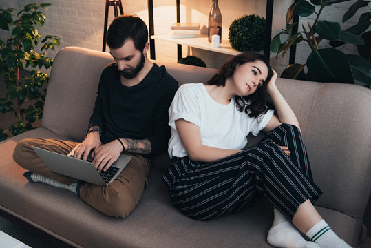 Bored Woman Sitting On Couch While Man Using Laptop In Living Room