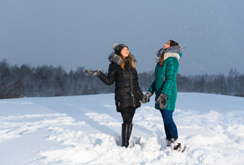 Fototapeta na wymiar Two happy Caucasian girls are holding hands and looking at the evening sky during snowfall in rural.