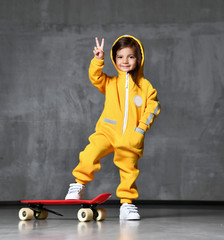 Young girl kid in yellow hooded jumpsuit with hood over her head and white sneakers stands on red skateboard and show peace sign © Dmitry Lobanov