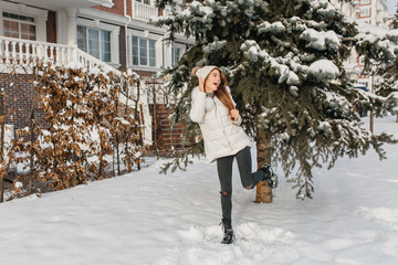 Charming tall girl in warm winter clothes having fun with snow on fir tree background in sunny...
