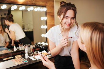 Cheerful young make up artist stand in front of blonde woman in beauty room and smile. She hold...