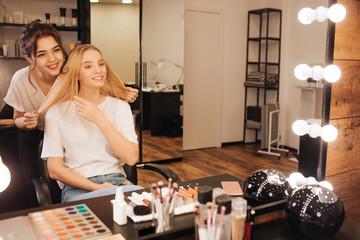 Nice cheerful young make up artist stand behind blonde model and smile to her in beauty room. she hold her hair and look into mirror. Client point on hair.