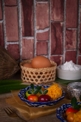 Obraz na płótnie Canvas Thai desserts on a plate of white and blue stripes placed on golden cloth and wood table there are pandan, water bowl, egg in basket, fork, flour, Similar object and coconut placed around.