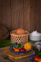 Thai desserts on a plate of white and blue stripes placed on golden cloth and wood table there are pandan, water bowl, egg in basket, fork, flour, Similar object and coconut placed around.