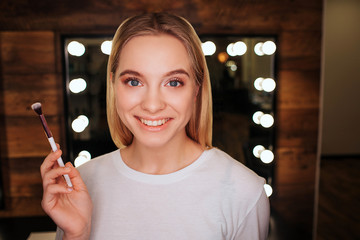 Nice young blonde woman look on camera and smile. She stand in beauty room. Woman hold brush for...