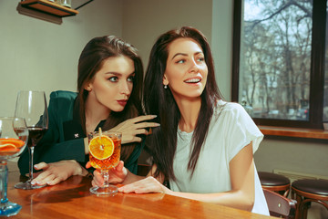 Female friends having a drinks at bar. They are sitting at a wooden table with cocktails. They are wearing casual clothes.