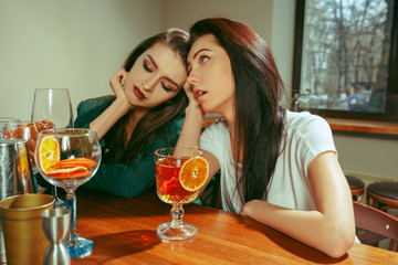 Female sad and tired friends having a drinks at bar. They are sitting at a wooden table with cocktails. They are wearing casual clothes.