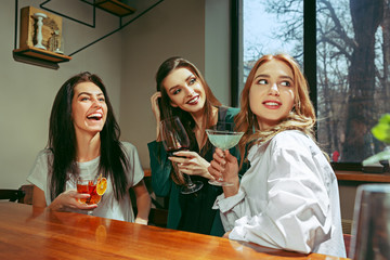 Female friends having a drinks at bar. They are sitting at a wooden table with cocktails. They are wearing casual clothes.