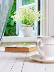 a cup of tea or coffee and a book on a white wooden antique retro table and a bouquet of lily-of-the-valley flowers on a window sill near an open window in a country house in the spring morning