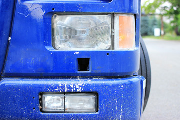 The rectangular headlight of an old blue truck, part of the bumper is stained with white paint.