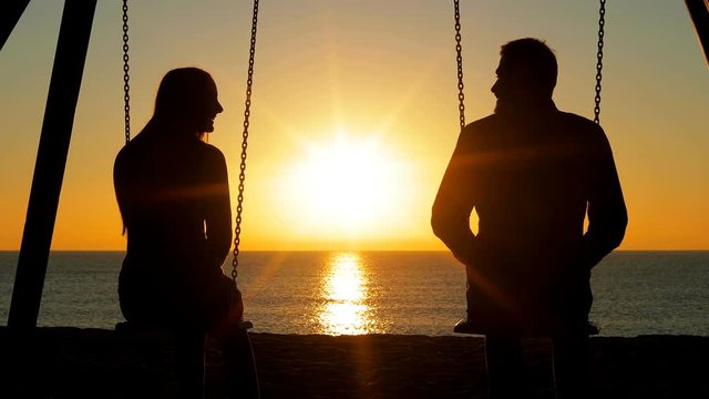 Back view silhouette of a happy couple swinging holding hands at sunrise on the beach