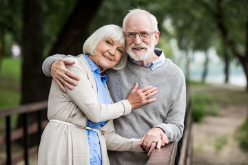 smiling senior couple hugging while standing by wooden bridge railing and looking at camera